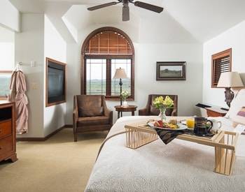 Wide angle view of the King Bed, Two Brown sitting chairs. Pass thru fireplace and a window with views of the mountain.